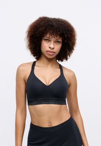 Črn bombažni športni nedrček z dizajnom v-izreza, širokimi naramnicami in elastičnim trakom z logotipom Calvin Klein. Gladek material, brez obloge.