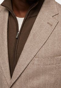 Blazer marrón con patrón de espiga sobre un suéter marrón con cremallera, que presenta una textura suave y un ajuste entallado, con detalles de cremallera visibles.