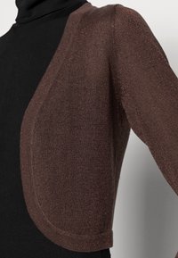 Zbliżenie na brązowy metaliczny krótki kardigan noszony na czarnym swetrze z golfem przez osobę.