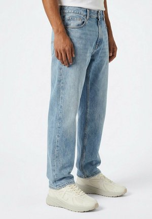 Persoon die lichtblauwe rechte jeans en off-white sportschoenen draagt, staand tegen een effen witte achtergrond.