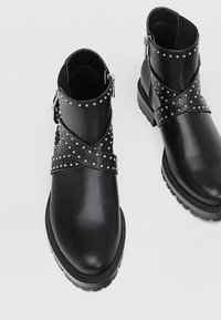 Bottines en cuir noir avec des sangles cloutées, des semelles en caoutchouc chunky et un design angulaire. Le détail zippé sur le côté améliore l'utilité.