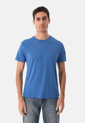 Uomo in piedi, rivolto verso l'obiettivo, indossa una maglietta blu semplice con un piccolo logo rosso e nero sul lato sinistro del petto e jeans blu.