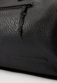 Čierna textúrovaná syntetická kožená taška s uzatváraním na zips a čiernym ťahákom, na ktorom je embosovaný logo nášivka s nápisom "ARMANI EXCHANGE."