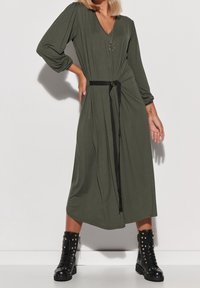 Robe longue vert olive avec col en V, manches longues, ceinture noire nouée à la taille et bas évasé, associée à des bottes de combat noires cloutées.