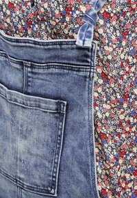 Les salopettes en denim présentent une poche avant, dans un lavage bleu clair avec une texture légèrement usée. L'arrière-plan montre une chemise à motifs floraux.