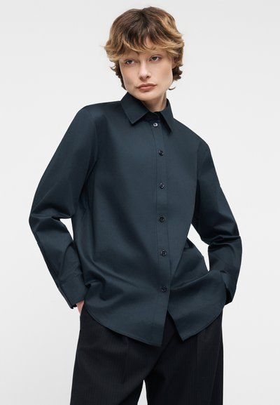 Άτομο με κοντά κυματιστά μαλλιά που φορεί σκούρο μακρυμάνικο πουκάμισο με κουμπιά και μαύρο παντελόνι, με τα χέρια στις τσέπες, μπροστά σε απλό φόντο.