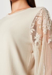 Beżowy sweter z miękkiego materiału, z przezroczystymi koronkowymi rękawami w kształcie bufek ozdobionymi kwiatowymi wzorami, tworzący kontrast w teksturze i wzornictwie.