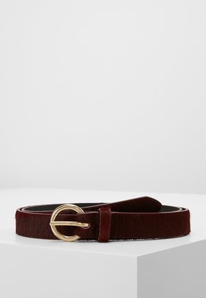 Ceinture en cuir marron foncé avec boucle ronde en métal doré, enroulée sur une surface blanche sur un fond gris clair.