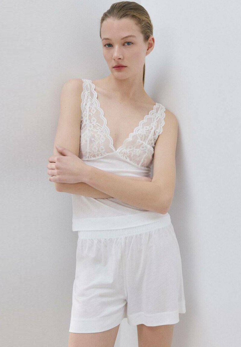 Mlada žena s plavom kosom koja je zavezana, nosi bijelu kratku majicu s čipkastim obrubom i odgovarajuće bijele kratke hlače, stoji s prekriženim rukama naslonjena na zid.