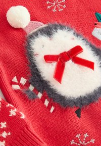 Rotes gestricktes Kleidungsstück mit einem Pinguin-Design, das weiße, flauschige Akzente, einen rosa Hut, eine rote Schleife und ein Zuckerstangenmuster aufweist. Weiche Textur sichtbar.