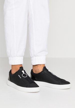 Føtter iført sorte lave sneakers med hvide såler og frontlynlås, kombineret med hvide manchetbukser mod en ensfarvet baggrund.