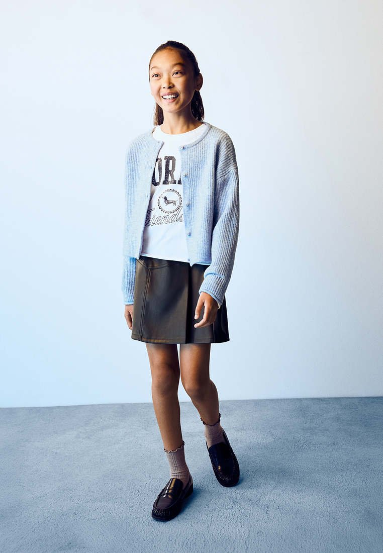 Jeune fille debout, portant un cardigan bleu clair, un t-shirt graphique blanc, une jupe foncée, des chaussettes beiges et des mocassins noirs, souriante et regardant devant elle.