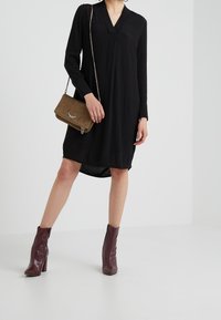 Vestido negro de manga larga con escote en V, que presenta un corte relajado y un dobladillo asimétrico. Combinado con un bolso cruzado de ante marrón y botas de tobillo burdeos.