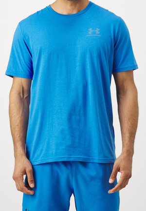 Mężczyzna ubrany w jaskrawo niebieską, sportową koszulkę z krótkim rękawem marki Under Armour oraz dopasowane niebieskie spodenki na jednolitym jasnym tle.