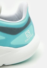 Turkos träningsskor med en slät, matt yta, med en vit dämpad sula och en rund häl. Visar "Salomon"-logotypen i vitt.