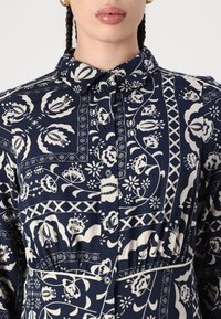 Camicia con bottoni blu navy con motivi floreali avorio, dotata di colletto, vita arricciata e dettagli geometrici.