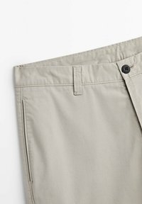 Gros plan sur la ceinture d’un pantalon beige clair avec passants de ceinture, poche avant et fermeture à bouton noir sur fond blanc.
