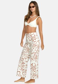 Weit geschnittene Hosen mit einem cremefarbenen Hintergrund, der mit großen roten Blumenmustern verziert ist. Strukturierter Stoff mit einer hohen Taille und einem Gürtel-Detail.