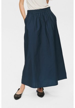 Dunkelblaue, knöchellange Rock mit elastischem Bund und seitlichen Taschen, aus glattem, leichtem Stoff. Flache, gewebte schwarze Schuhe.