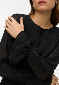 Melna, ar ziedu rakstu rakstīta blūze ar teksturētu audumu, garām caurspīdīgām zvana piedurknēm un apaļu izgriezumu, izceļot mīkstu audumu un elegantas dizaina detaļas.