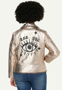 Metallisch goldene Lederjacke mit schwarzem Augen-Graphic und "SEE YOU"-Text auf dem Rücken. Mit Nietenabschluss und Kettenakzent am Kragen.