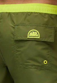 Shorts vert olive avec une ceinture élastique vert clair, une poche arrière avec un logo à bord jaune et un détail en œillet sur le côté.