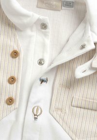 Polo blanc avec des rayures verticales beiges, des boutons en bois, une fermeture à pression argentée, et des motifs brodés de ballon et de bobine.