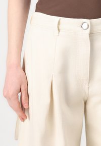 Crème-kleurige broek van textuurstof, met een passend middel met een zilveren knop en voorplooien voor extra detail.