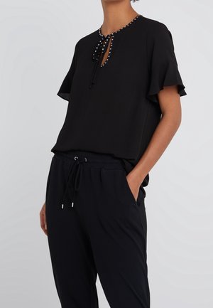Mujer con blusa negra con escote adornado con perlas y mangas acampanadas, combinada con pantalones negros con cordón, mano en el bolsillo.