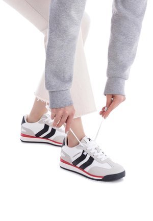 Zapatillas deportivas con un upper gris y blanco, rayas negras, acentos rojos y suela texturizada. Se están atando los cordones en el zapato izquierdo.