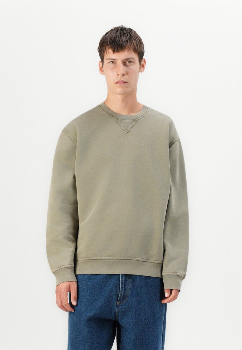 Sudadera verde olivo con cuello redondo y puños acanalados. Tejido suave, corte holgado, con un sutil detalle de costura en el hombro.