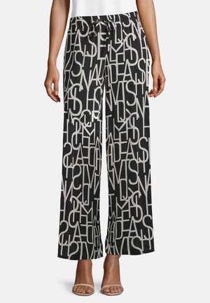 Mustat leveät housut, joissa on päällekkäisiä suuria valkoisia kirjaimia, elastinen vyötärönyöri, käytettynä valkoisten sandaalien ja valkoisen paidan kanssa.