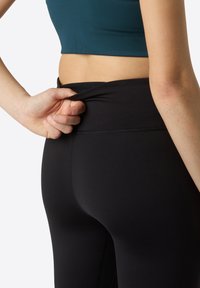 Leggings noirs avec une taille haute, en tissu élastique lisse. Une main tire sur la taille pour ajuster le fit.