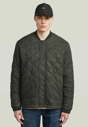 Junger Mann trägt eine schwarze gesteppte Jacke, blaue Jeans und eine schwarze Baseballkappe vor einem einfarbigen hellen Hintergrund.