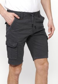 Man med mörkgrå cargo-shorts med dragsko i midjan och sidofickor, stående med ena handen i fickan.