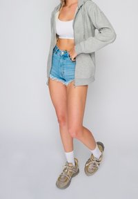 Felpa zip-up grigia chiara con cappuccio, top corto bianco, shorts di jeans strappati, e sneaker beige-grigie con dettagli gialli e calze bianche.