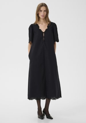 Žena v čiernych midi šatách so zaoblenými okrajmi, krátkymi naberanými rukávmi, čiernych pančuchách a čiernych lodičkách s ostrou špičkou, stojaca s rukami v kapse.