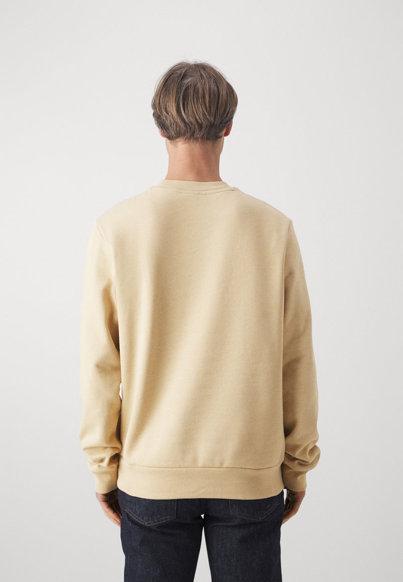 Beiger Pullover mit Rundhalsausschnitt, gerippten Bündchen und Saum sowie einer glatten Oberfläche. Wird über dunklen Jeans getragen und hat lange Ärmel.