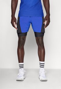 Pantalones cortos deportivos azules con detalles en negro, con un diseño ajustado y una textura de malla, combinados con zapatillas deportivas blancas y calcetines rayados.