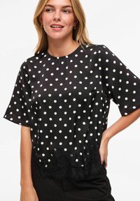 Blouse noire à pois avec un col rond, des manches courtes et un ourlet orné de dentelle. Tissu lisse, grands pois blancs et détails délicats en dentelle.