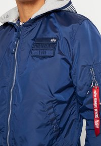 Marinblå bomberjacka med en grå fleecehuva, dragkedja framtill, broderad patch och en röd "Remove Before Flight"-tagg på ärmen.