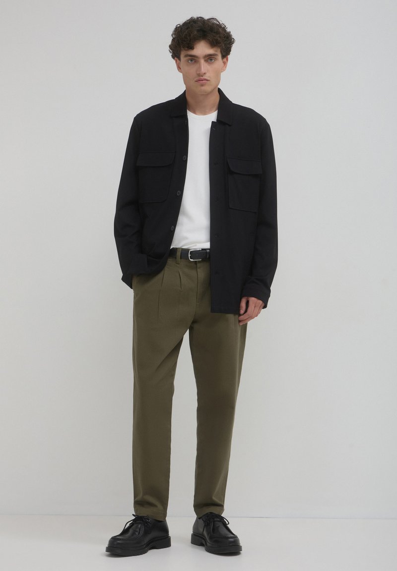 Schwarze Knopfhemd mit zwei Brusttaschen, darunter ein weißes T-Shirt, olivgrüne Hose und schwarze Schuhe. Glatte Texturen und ein einfaches Design.