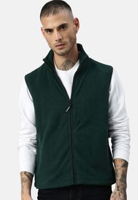 Dunkelgrüne Fleece-Weste mit hohem Kragen und Frontreißverschluss, getragen über einem weißen Langarmshirt. Hat eine glatte Textur und ein lässiges Design.