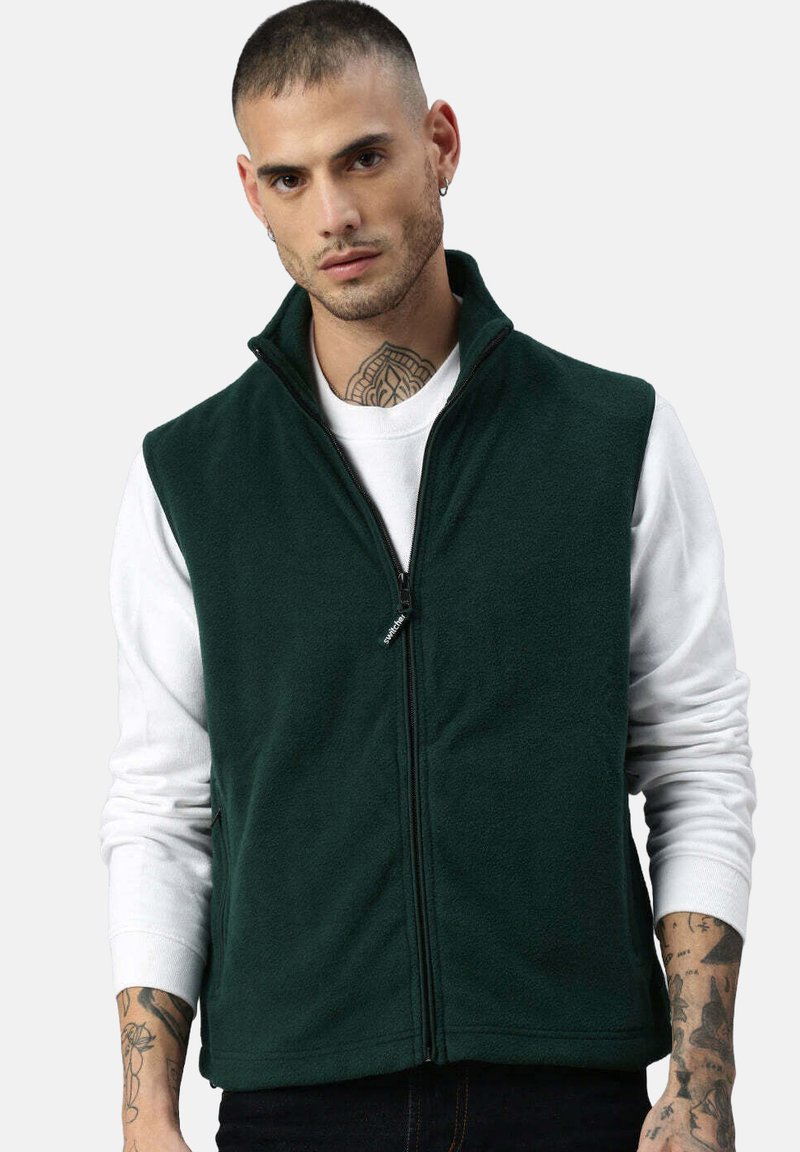 Dunkelgrüne Fleece-Weste mit hohem Kragen und Frontreißverschluss, getragen über einem weißen Langarmshirt. Hat eine glatte Textur und ein lässiges Design.