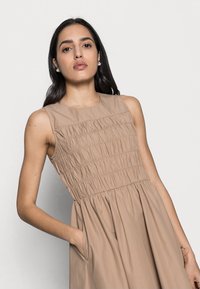 Beiges Kleid mit einem taillierten, gerafften Oberteil und einem schwingenden Rock. Es hat ein ärmelloses Design und seitliche Taschen für zusätzlichen Komfort.