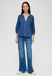Blouse légère et bleue avec un décolleté en V et des détails froncés. Associée à un jean en denim évasé et des baskets couleur crème.