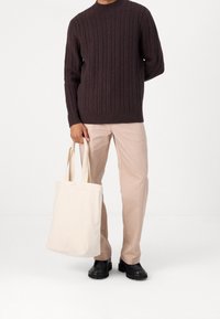 Pull en maille torsadée marron foncé, pantalon beige coupe droite et sac fourre-tout beige clair avec anses plates ; des chaussures noires épaisses complètent le look.