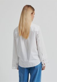 Chemise blanche en coton à manches longues avec des découpes aux poignets, portée avec un jean en denim bleu.