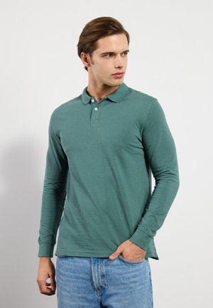 Långärmad grön pikétröja i bomull; har en tre-knappsknäppning, ribbade ärmslut och en något rundad nederkant. Bärs tillsammans med jeans.