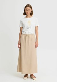 T-shirt blanc avec un graphique "WILD LIFE" et jupe maxi beige avec une taille élastique. Modèle portant des chaussures de couleur claire.